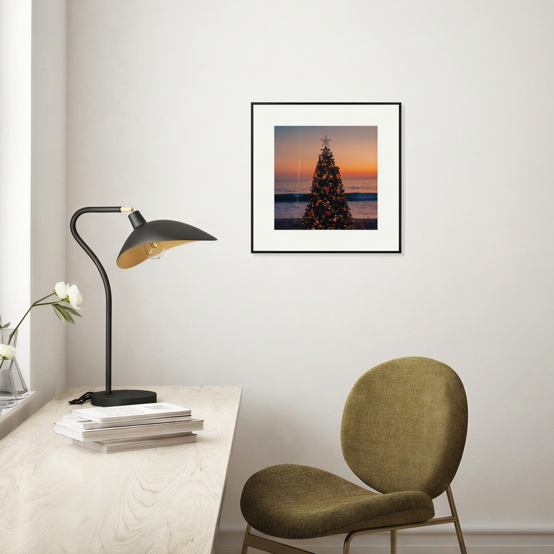 Framed artwork of a Christmas tree against a sunset sky on a wall above a desk with a lamp and chair.