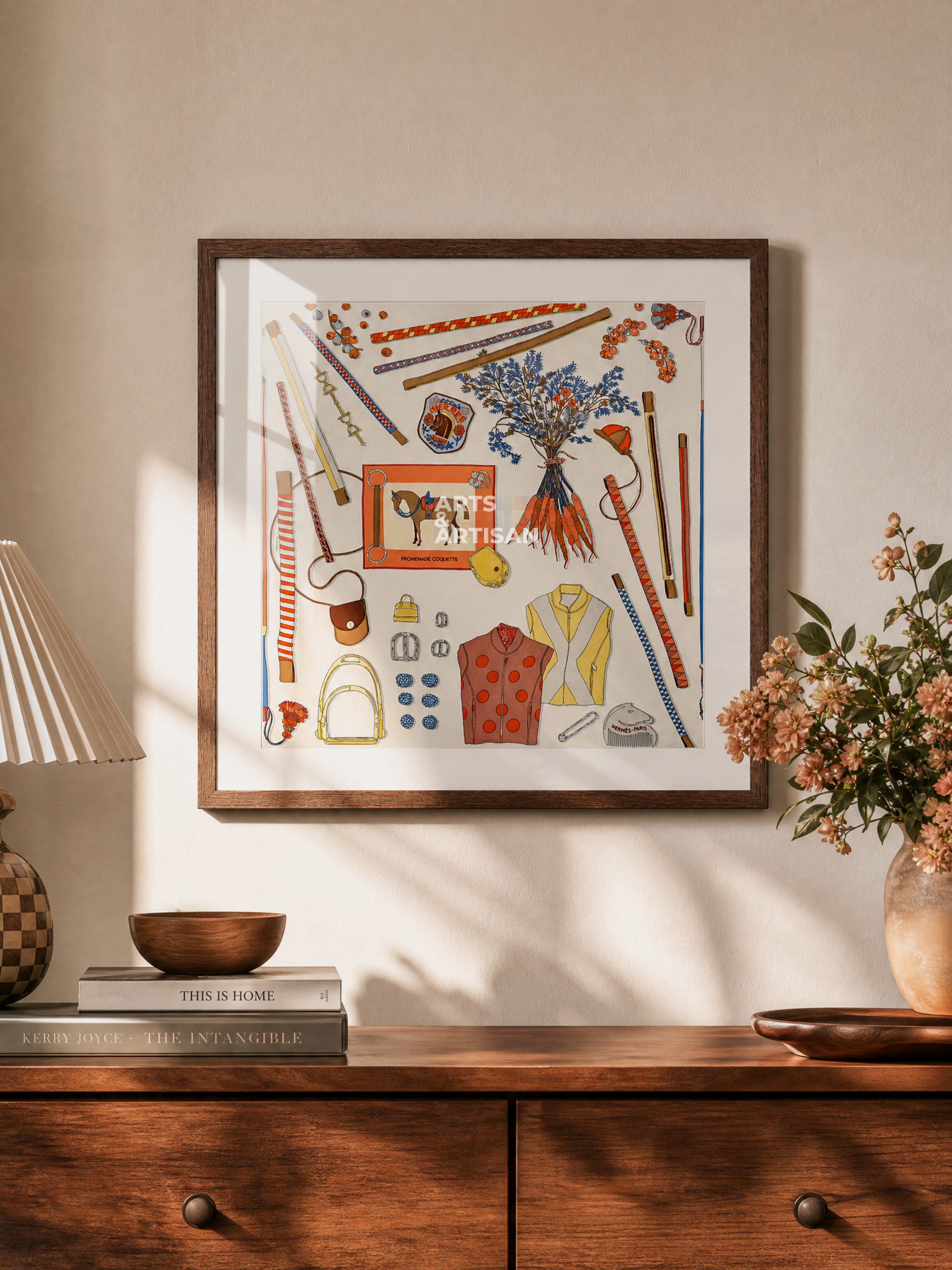 A framed Hermes Scarf 70 featuring a colorful collage of sewing and fashion-themed illustrations, displayed on a beige wall above a wooden console table with a lamp, books, and flowers.