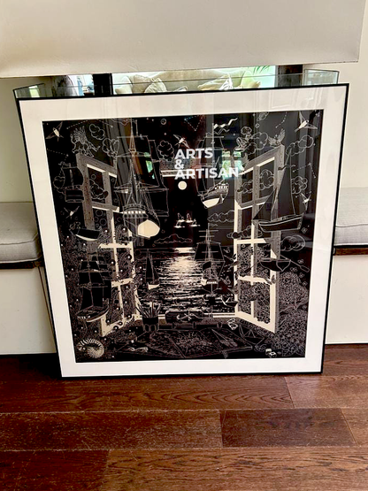 A framed black Hermes scarf leaning against a white wall on a dark wood floor. The print features an abstract, high-contrast architectural illustration with the text 'ARTS & ARTISAN' in the center.