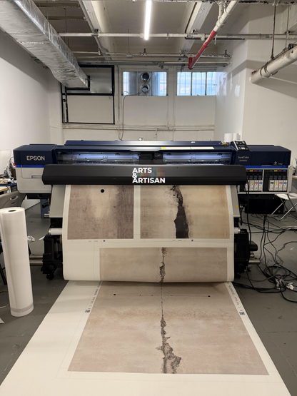 An Epson Arts Artisan large-format UV printer in a workshop setting, printing a textured beige and brown fabric or wallpaper sample with a distressed, cracked pattern onto a wide roll of material.