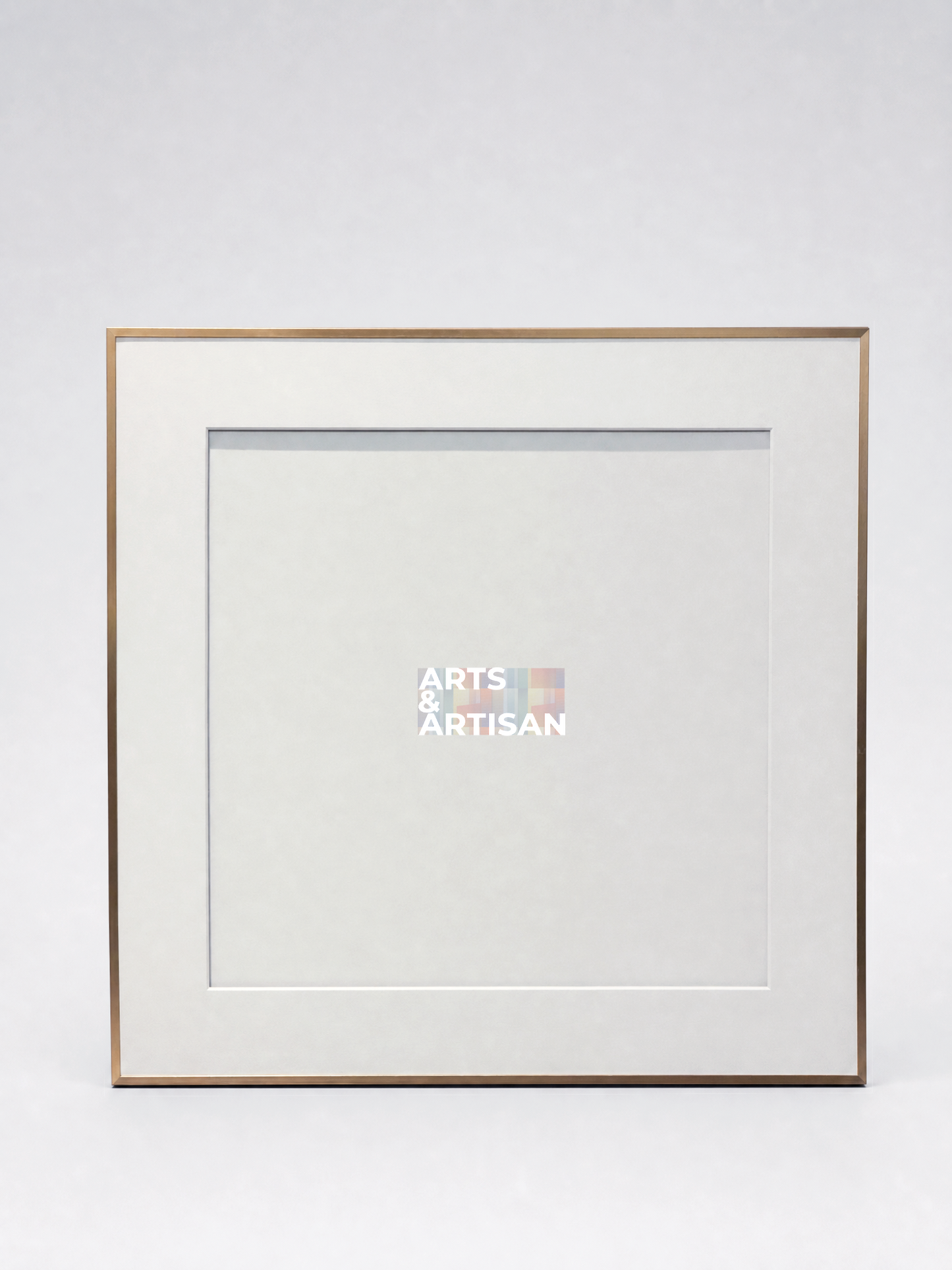 A square picture frame with a white mat and a thin gold-toned metal border, shown against a light gray studio background. A small Arts & Artisan logo is visible in the center of the mat.