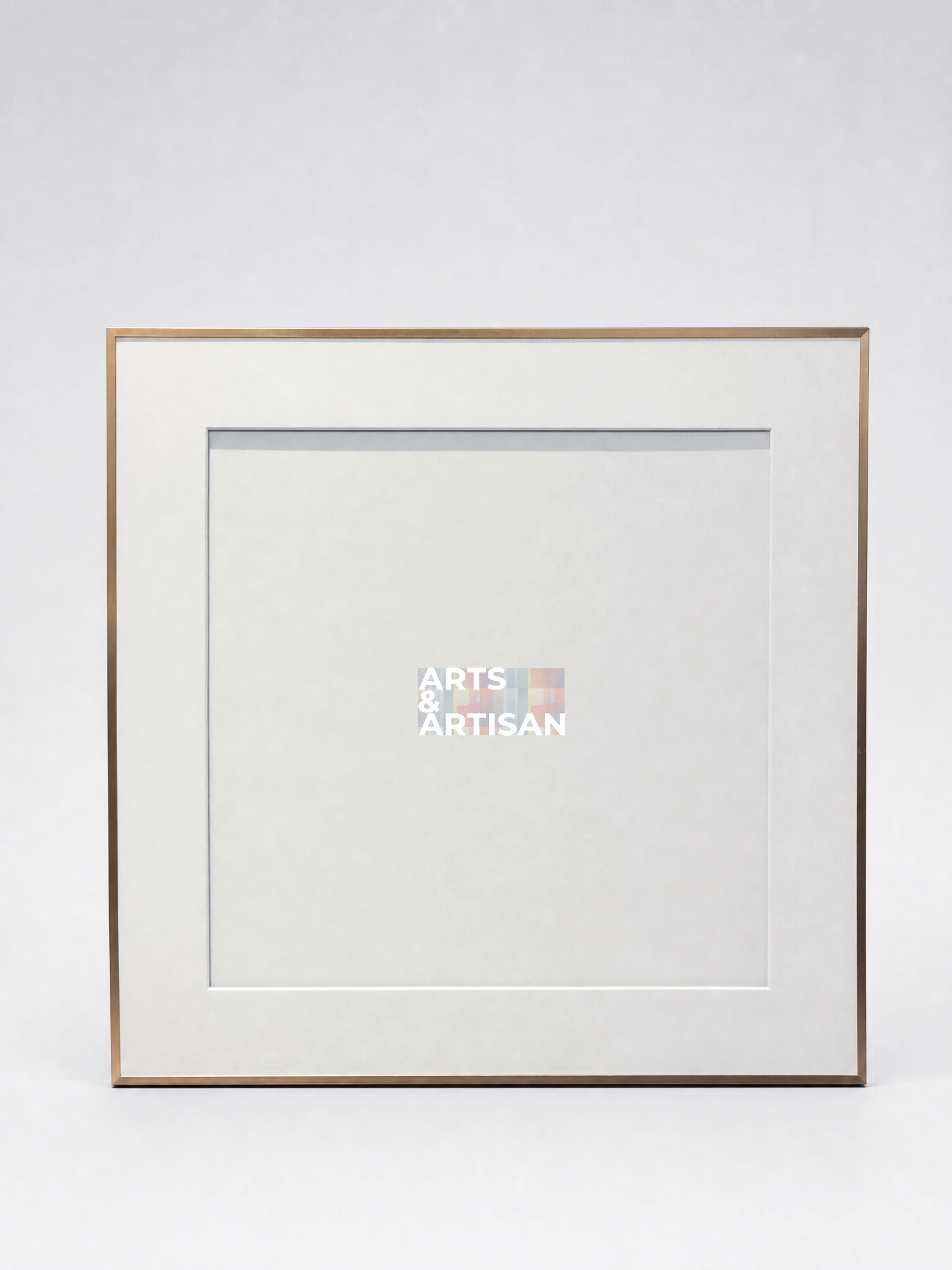 A square picture frame with a white mat and a thin gold-toned metal border, shown against a light gray studio background. A small Arts & Artisan logo is visible in the center of the mat.