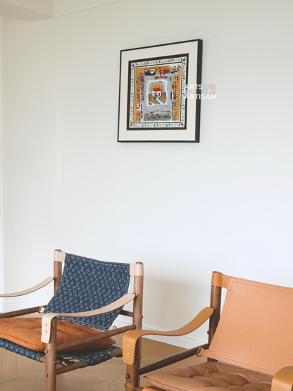 A framed scarf 45 frame with a colorful architectural scene hangs on a white wall above two wooden chairs, one with a blue patterned fabric seat and one with a tan leather seat.